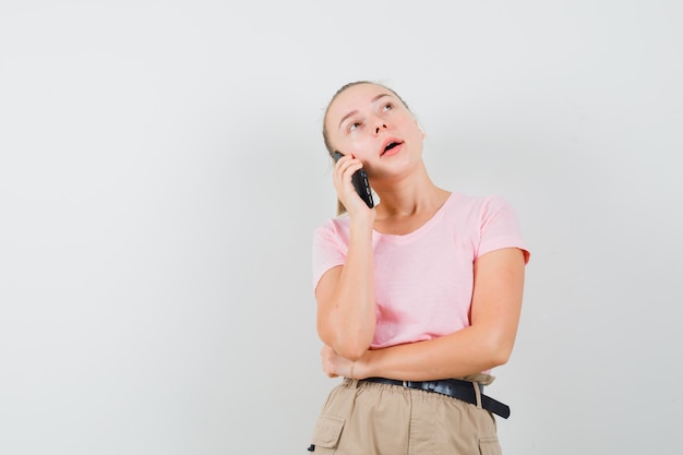 Блондинка разговаривает по мобильному телефону в футболке, штанах и задумчиво, вид спереди.