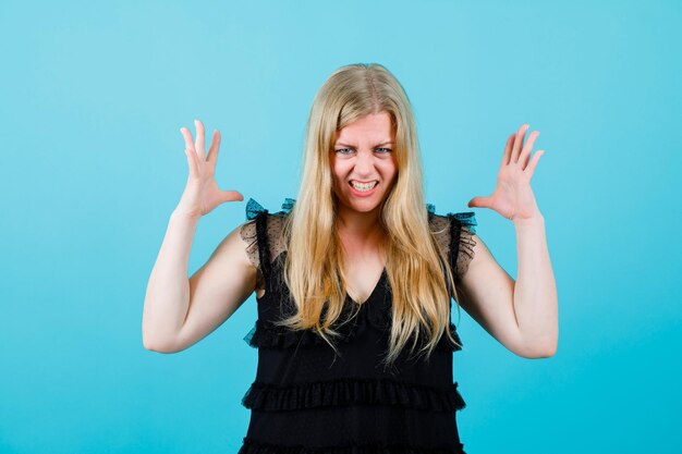 Blonde girl with angry mimciry is raising up her hands on blue background