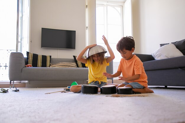 Blonde girl with bowl on head playing with friend. Cheerful little boy knocking on pans. Two happy children sitting on floor and having fun together in living room. Childhood, holiday and home concept