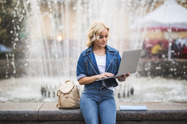 Blonde girl woman student works on her laptop computer near fountain in the city in the day