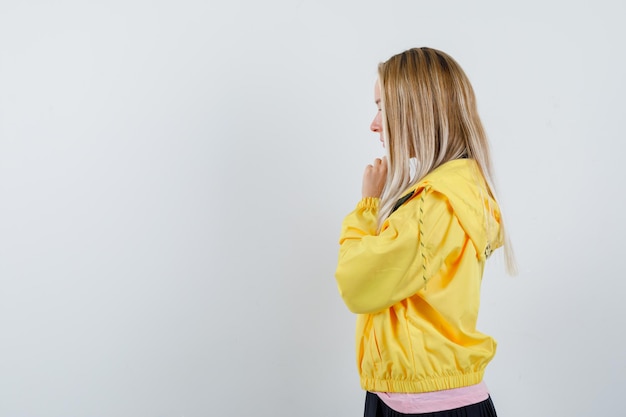 Ragazza bionda in giacca gialla che stringe le mani in un gesto di preghiera e sembra speranzosa.