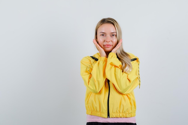 Blonde girl in yellow jacket holding hands near cheeks and looking delicate