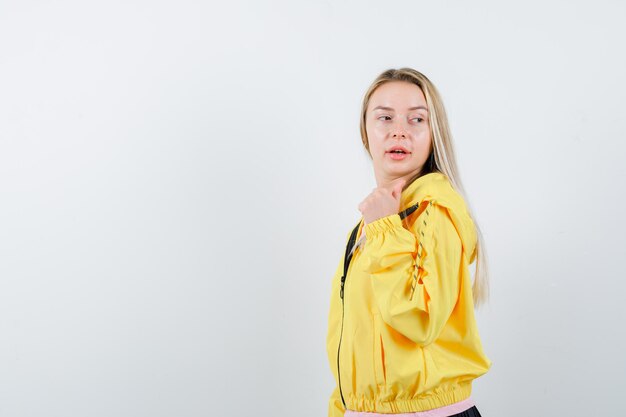 Блондинка в желтой куртке, указывая большим пальцем назад, глядя через плечо и выглядя заманчиво.