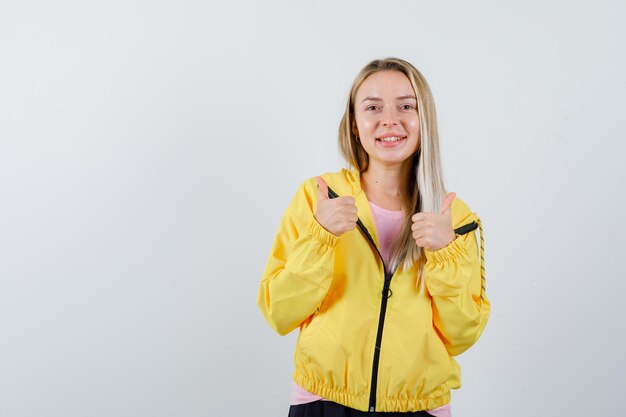 Блондинка в желтой куртке показывает двойные пальцы вверх и выглядит счастливой