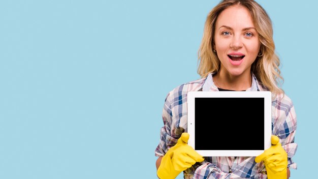 Blonde hair young woman with mouth open wearing gloves holding digital tablet in front of blue backdrop