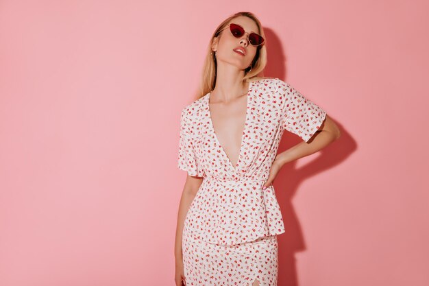 Blonde haired woman in round gold earrings red stylish sunglasses and white cool clothes looking into camera on pink background