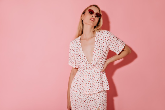 Blonde haired woman in round gold earrings red stylish sunglasses and white cool clothes looking into camera on pink background