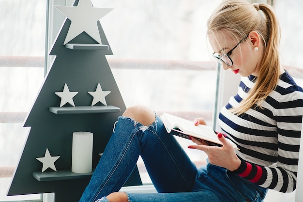 Blonde lady in black glasses reads book sitting before wooden Christmas tree on balcony