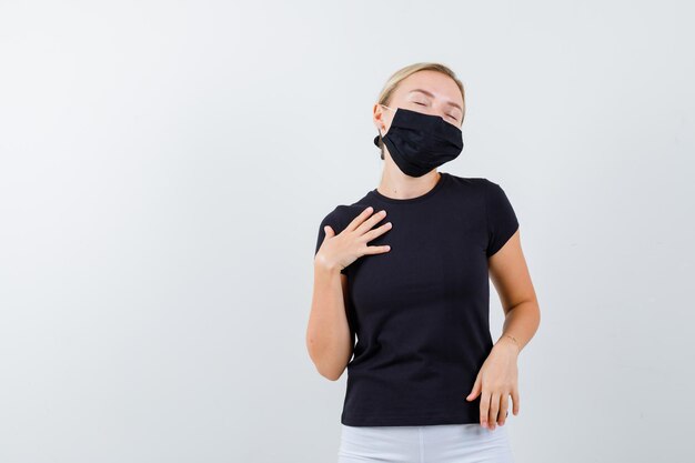 黒のTシャツ、孤立した胸に手を保持している黒のマスクのブロンドの女性