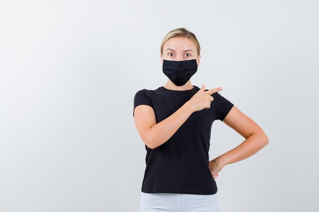 Blonde lady in black t-shirt, black mask pointing to the right side isolated