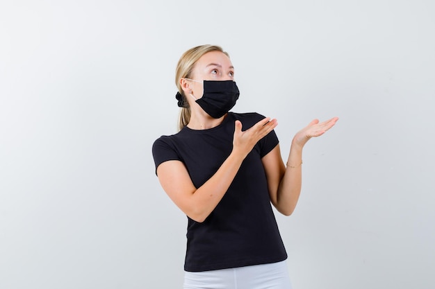 Blonde lady in black t-shirt, black mask pretending to show something isolated