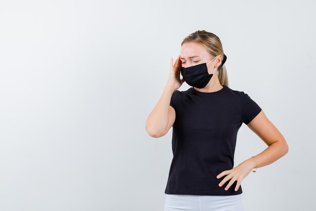 Blonde lady in black t-shirt, black mask rubbing temples and looking painful