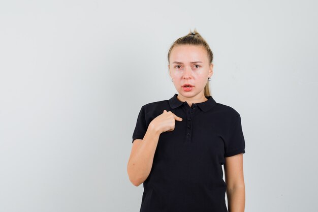 Blonde lady in black t-shirt pointing at herself and looking puzzled , front view.