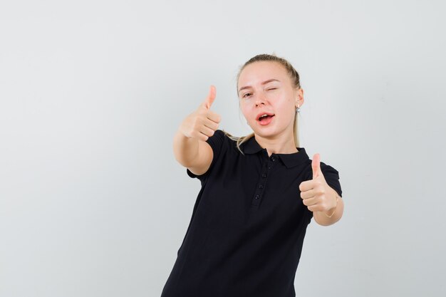 Blonde lady in black t-shirt showing double thumbs up, winking eye and looking confident , front view.