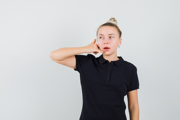 Blonde lady in black t-shirt showing phone gesture while looking aside , front view.