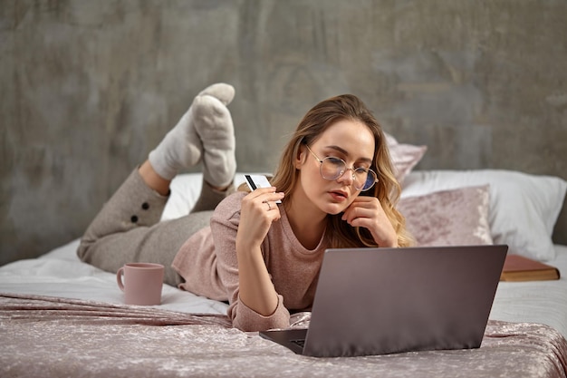 Blonde lady blogger in glasses casual clothing holding white plastic card lying on bed with laptop b...