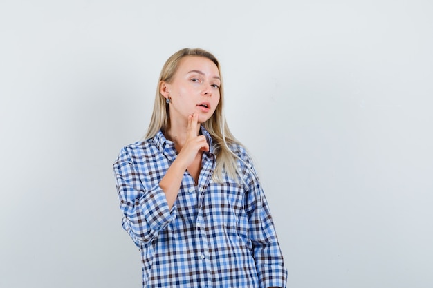 Blonde lady in casual shirt holding finger on chin and looking pensive