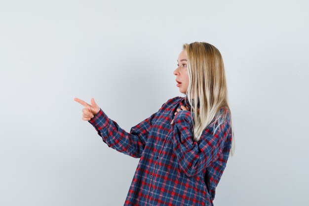 Blonde lady in casual shirt pointing away and looking wondered , front view.