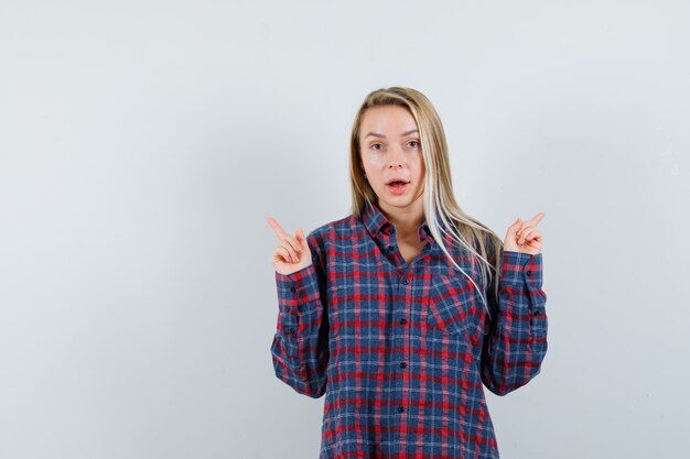 Blonde lady in casual shirt pointing up and looking surprised , front view.