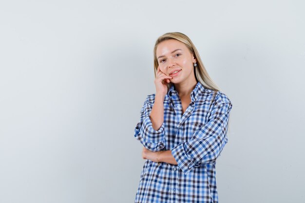 Blonde lady in checked shirt holding hand on cheek and looking glad