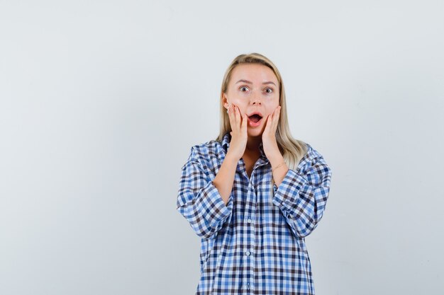 Blonde lady in checked shirt holding hands on cheeks and looking astonished