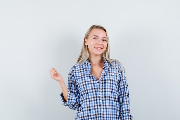 Blonde lady in checked shirt posing while raising hand and looking joyful