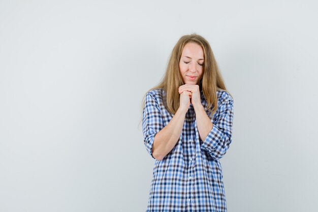 Blonde lady clasping hands in praying gesture in shirt and looking hopeful ,