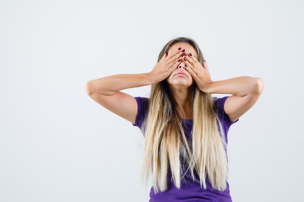 Blonde lady covering eyes with hands in violet t-shirt front view.