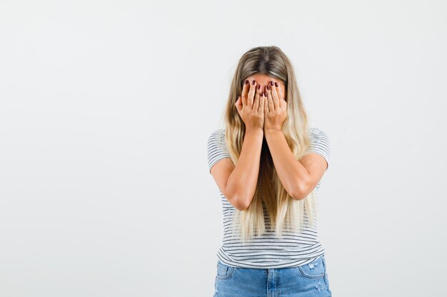 Blonde lady covering her face with hands in t-shirt and looking sad. front view.