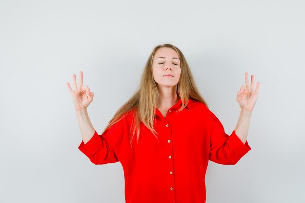 Blonde lady doing meditation with closed eyes in red shirt and looking relaxed.