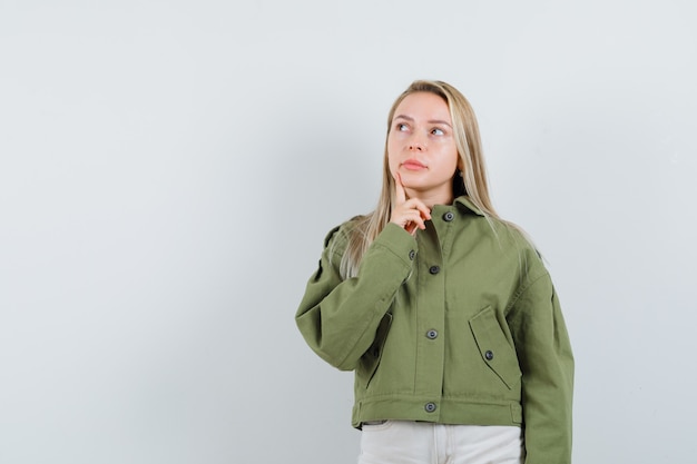 Blonde lady holding finger on chin in jacket, pants and looking pensive , front view.