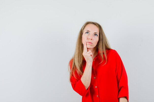 Blonde lady holding finger on chin in red shirt and looking pensive.