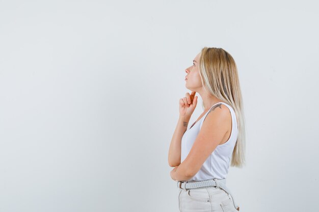 Blonde lady holding finger on chin in singlet, pants and looking thoughtful .