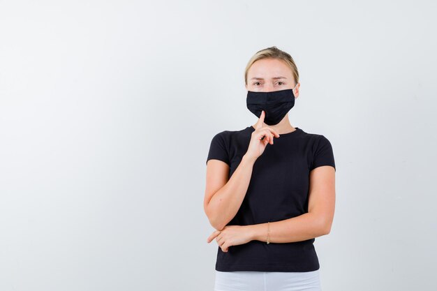 分離された黒いTシャツで彼女の医療マスクに指を保持しているブロンドの女性