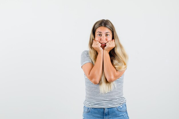 Blonde lady holding fists on her cheeks while listening in t-shirt and looking attentive , front view.