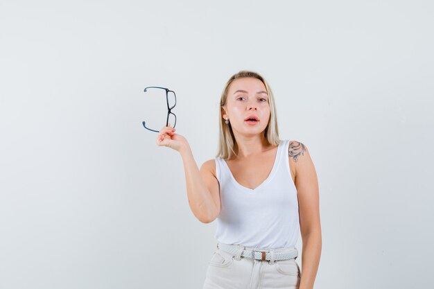 Blonde lady holding glasses in singlet, pants and looking pensive. front view.