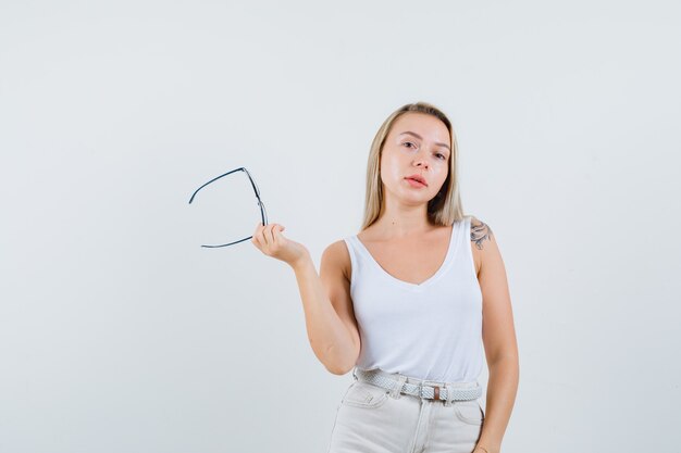 Blonde lady holding glasses in singlet, pants and looking sensible