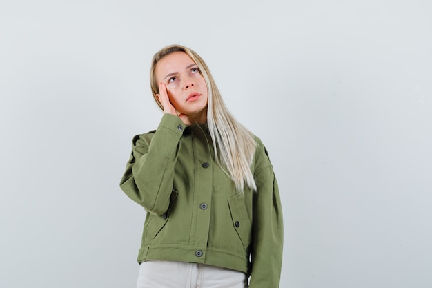 Blonde lady holding hand on cheek in jacket, pants and looking thoughtful , front view.