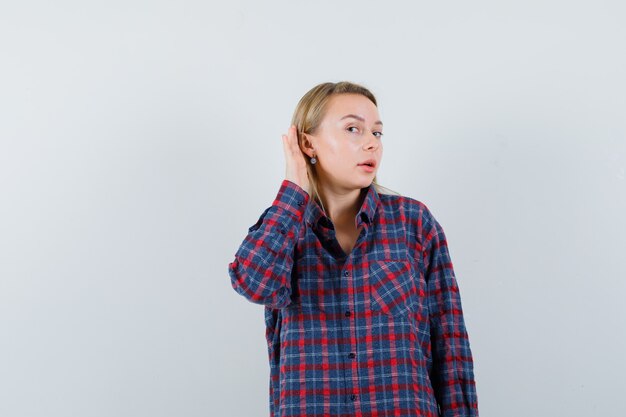 Blonde lady holding hand behind ear in casual shirt and looking curious. front view.