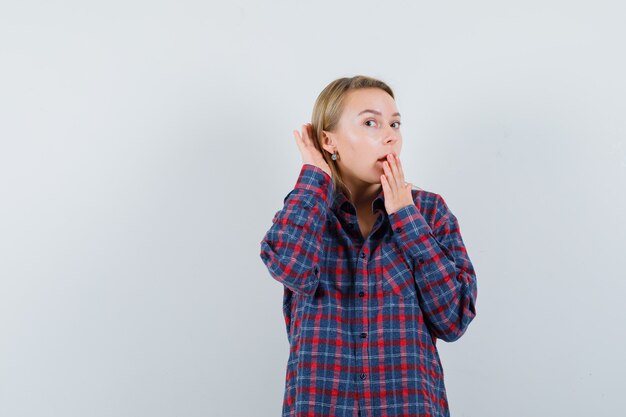 Blonde lady holding hand behind ear in casual shirt and looking surprised , front view.