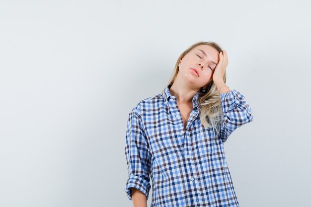 Blonde lady holding hand on head in casual shirt and looking sleepy