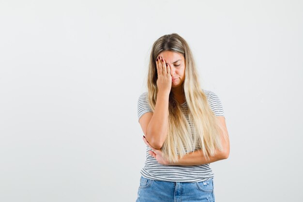 Blonde lady holding hand on her face in t-shirt and looking tired. front view.