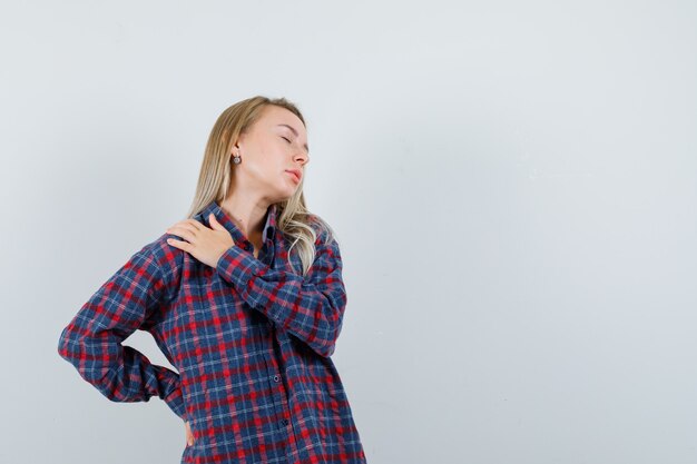 Blonde lady holding hand on shoulder in casual shirt and looking fatigued , front view.