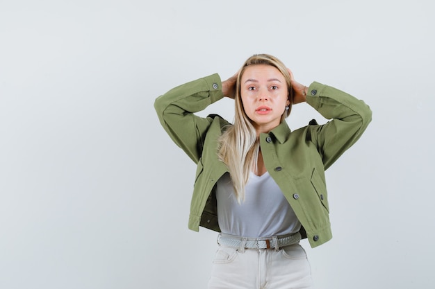 Blonde lady holding hands behind head in jacket, pants and looking forgetful , front view.