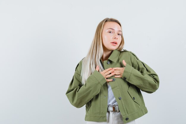 Blonde lady holding hands on heart in jacket, pants and looking pretty. front view.