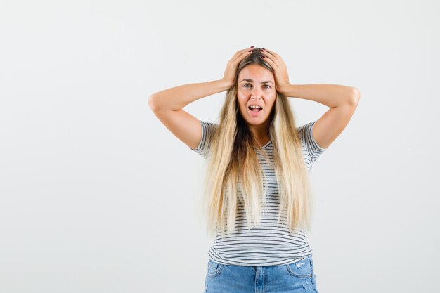 Blonde lady holding hands on her head in t-shirt and looking confused. front view.