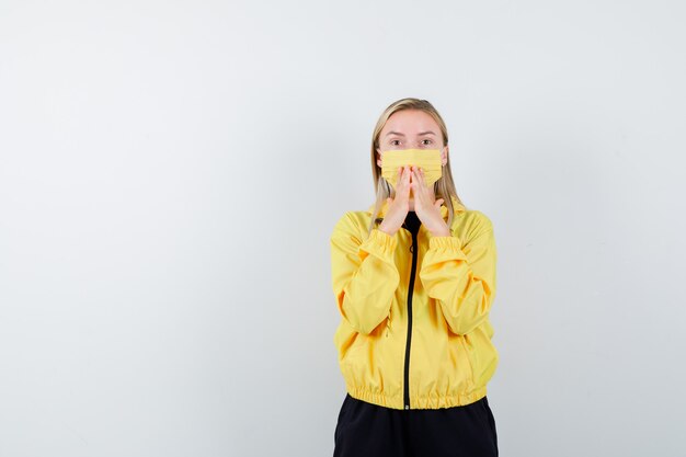 Free photo blonde lady holding hands on mouth in tracksuit, mask and looking anxious , front view.