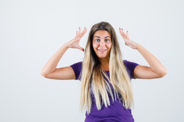 Blonde lady holding hands near head in violet t-shirt and looking happy. front view.