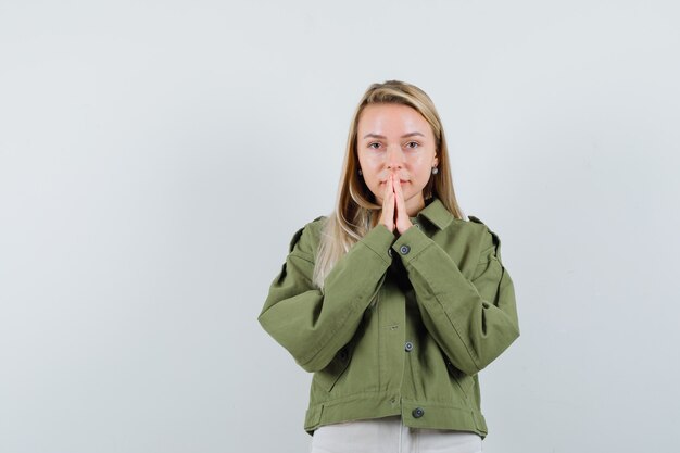 Blonde lady holding hands in praying gesture in jacket, pants and looking positive. front view.