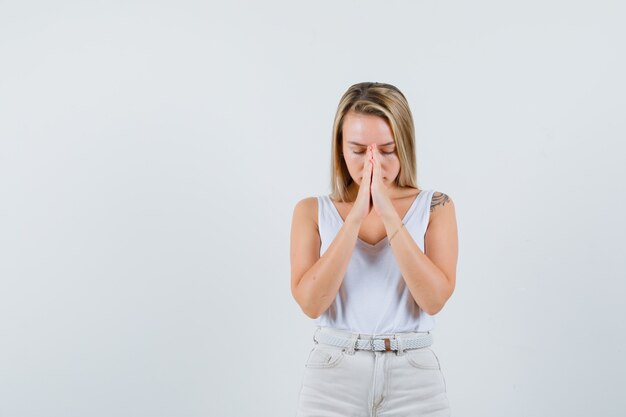 Blonde lady holding hands in praying gesture in singlet, pants and looking hopeful